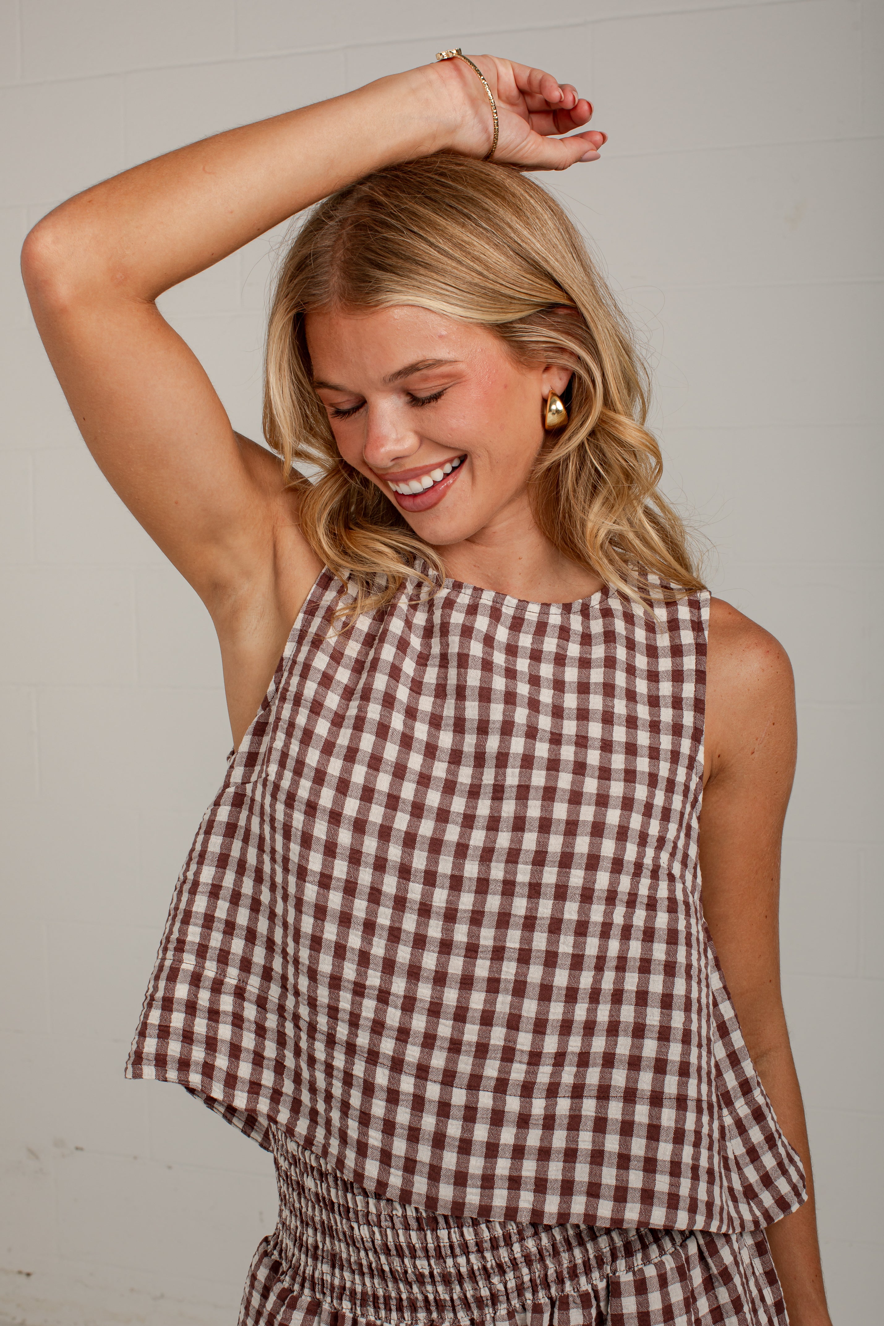 A woman wearing a brown and white gingham sleeveless top and matching bottoms, smiling with one arm raised against a plain light background.