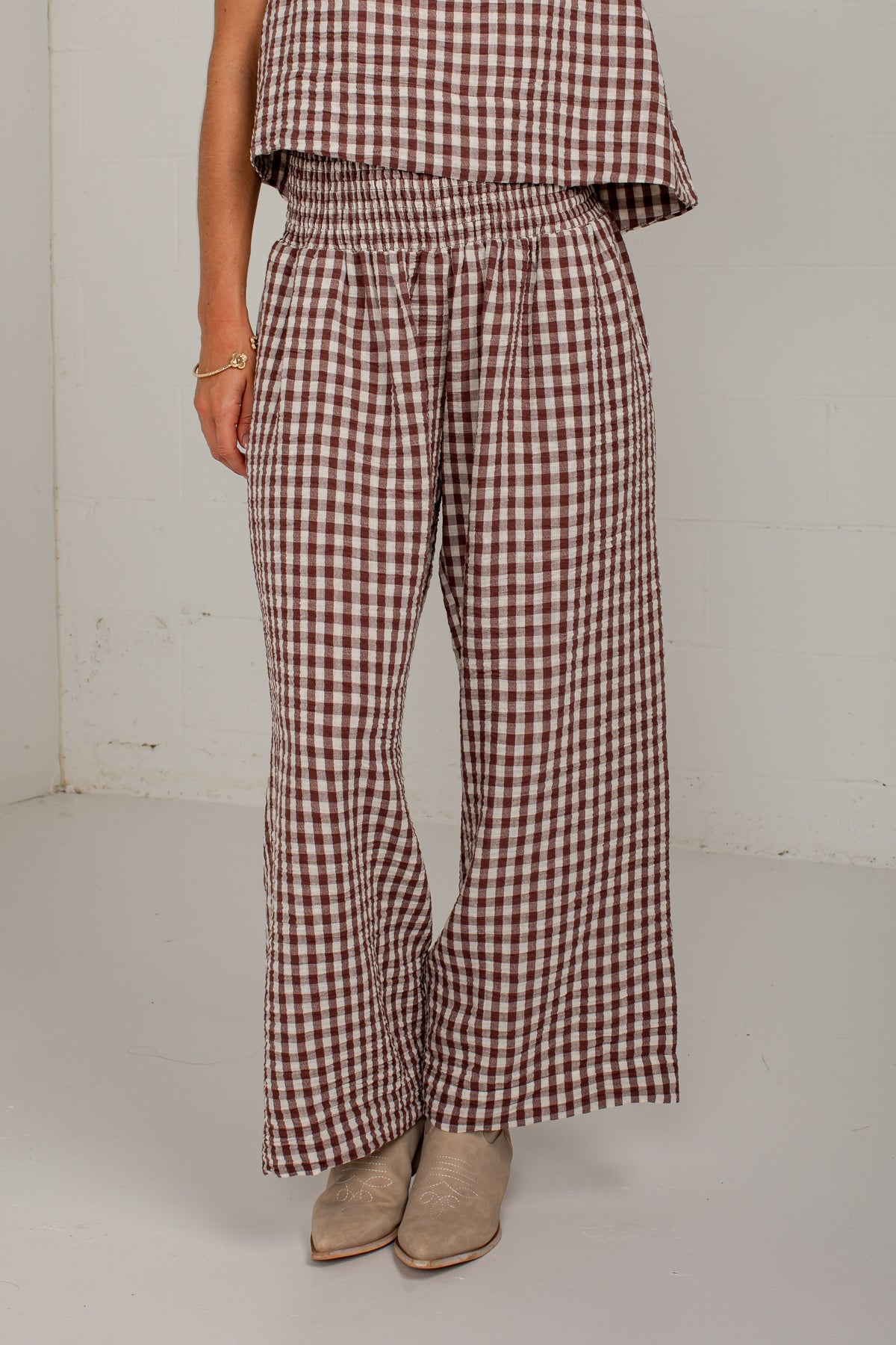 A person wearing brown and white gingham wide-leg pants with an elastic waistband, paired with beige ankle boots, photographed in a plain indoor setting.