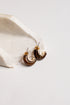 A pair of crescent-shaped hoop earrings with a glossy dark brown and gold finish, resting on a textured white stone prop against a clean white background.