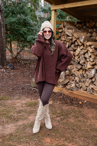 Woman in a brown sweater and white boots standing in front of stacked firewood.