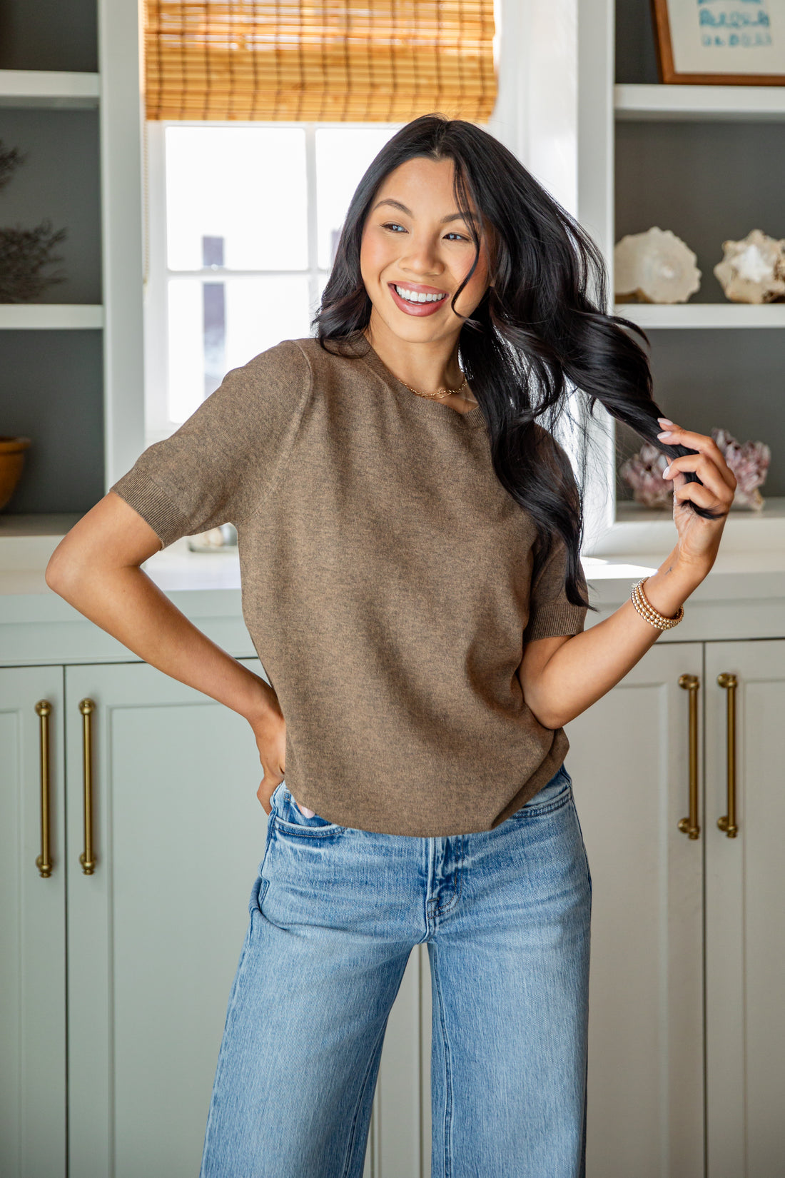 Woman in a brown sweater and blue jeans standing in a kitchen.