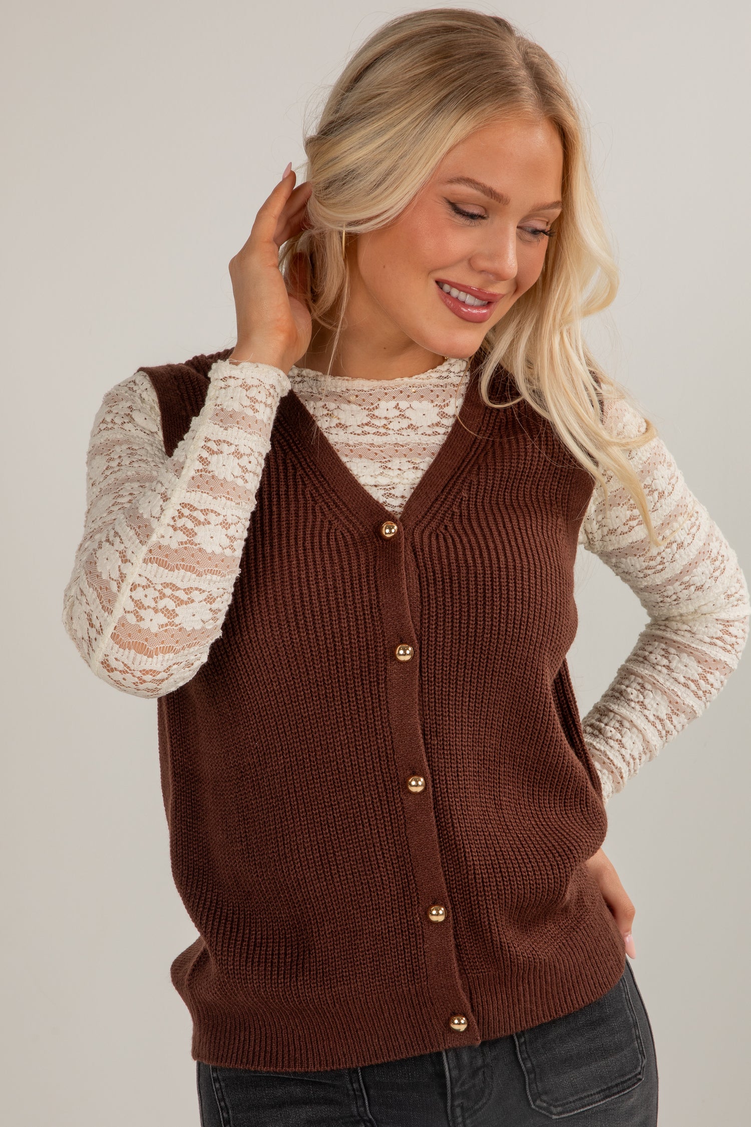 Woman wearing a brown button-up vest over a white lace top on a plain background