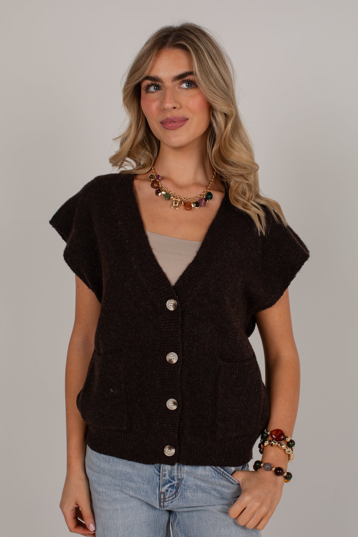 A woman modeling a dark brown short-sleeve knit cardigan with silver buttons, worn over a beige top and paired with light blue jeans, against a plain light gray background.