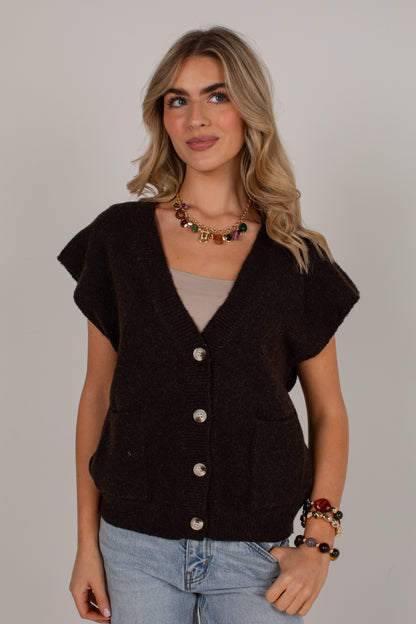 A woman modeling a dark brown short-sleeve knit cardigan with silver buttons, worn over a beige top and paired with light blue jeans, against a plain light gray background.