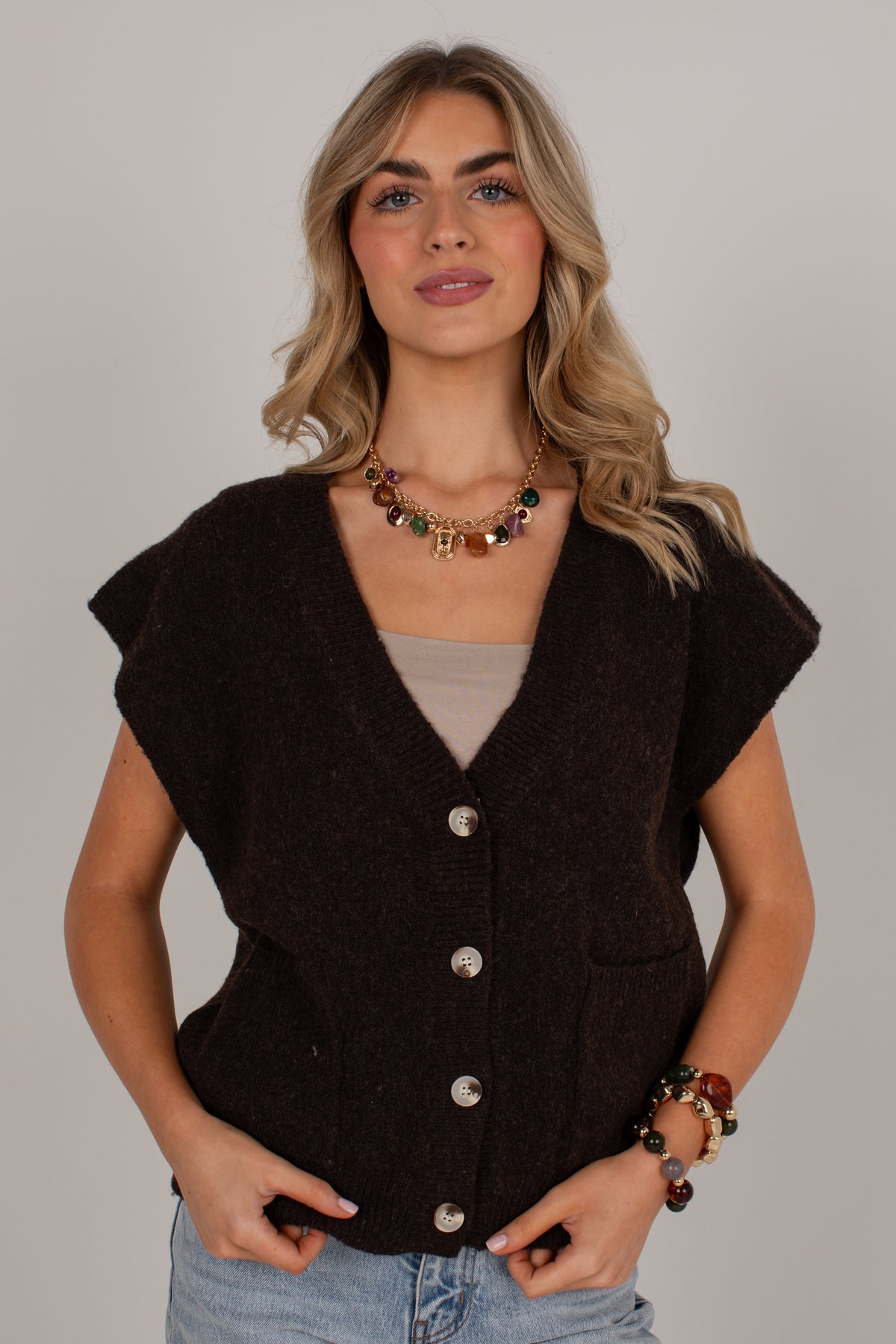 A woman modeling a dark brown sleeveless knit cardigan vest with silver buttons, worn over a beige top and blue jeans, against a plain light gray background.