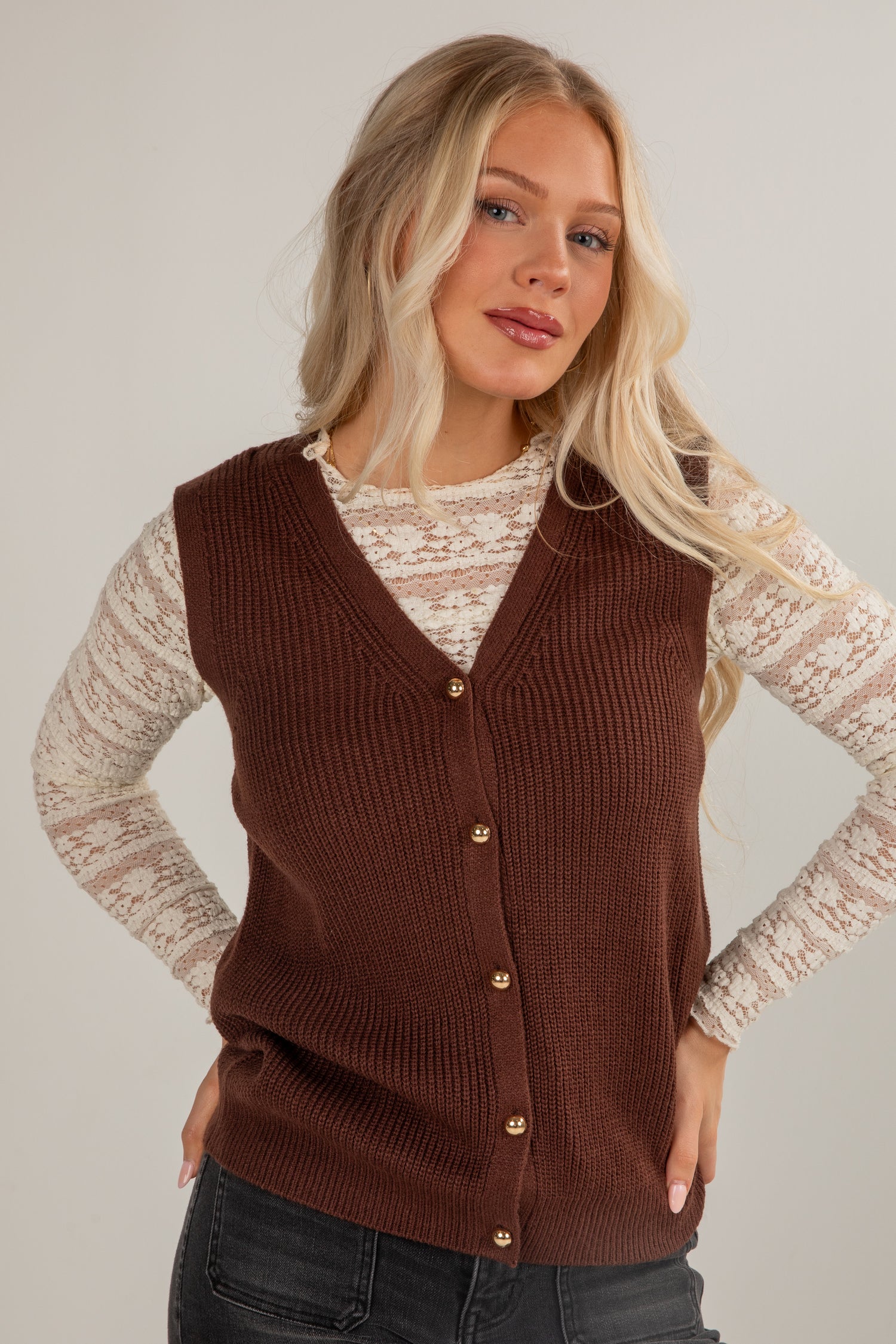 Woman wearing a brown knitted vest over a lace top against a plain background