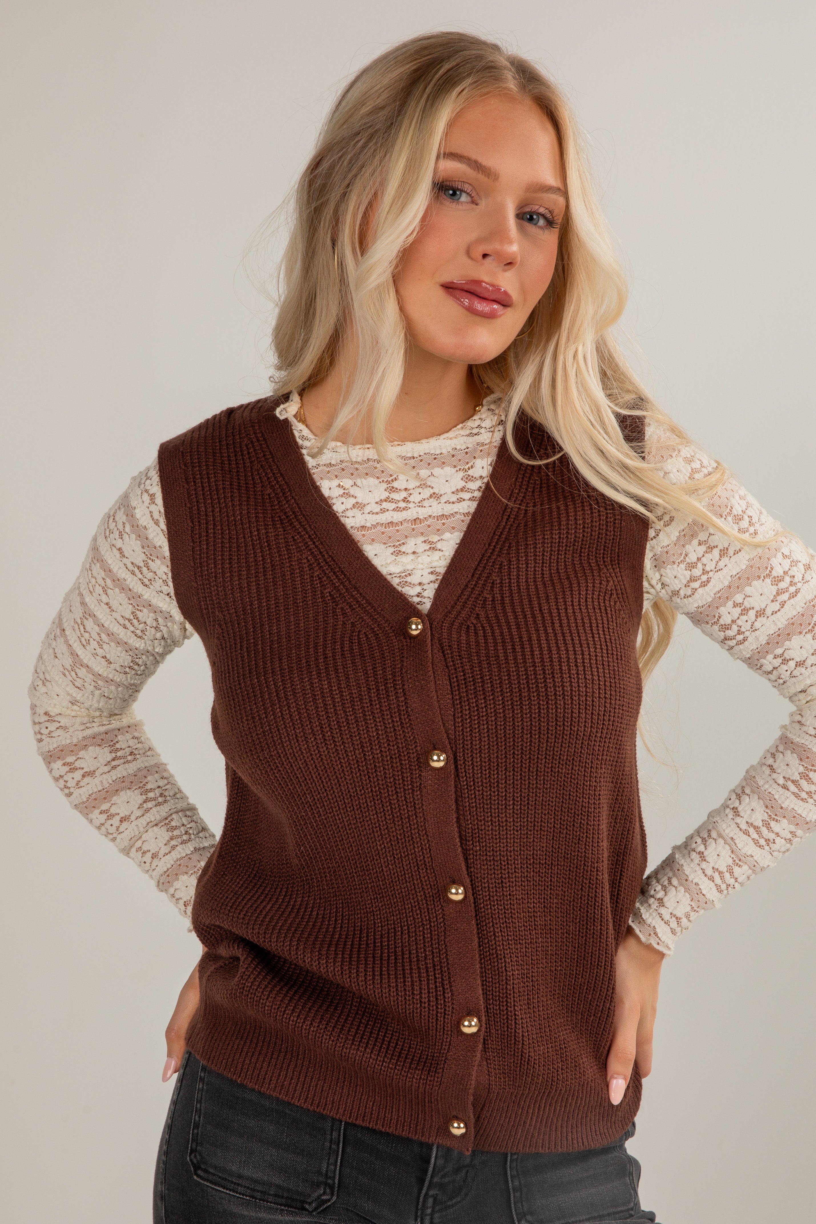 Woman wearing a brown knitted vest over a lace top against a plain background