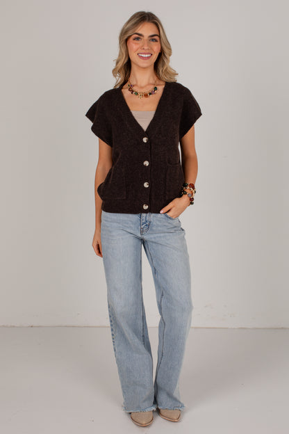 A woman modeling a dark brown short-sleeve knit cardigan vest with front buttons, worn over a light gray top and paired with light-wash blue jeans, against a plain light gray studio background.
