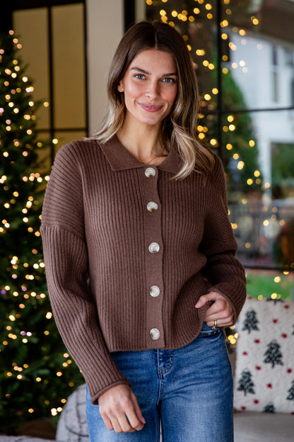Woman wearing a brown ribbed sweater with white buttons in a festive indoor setting.