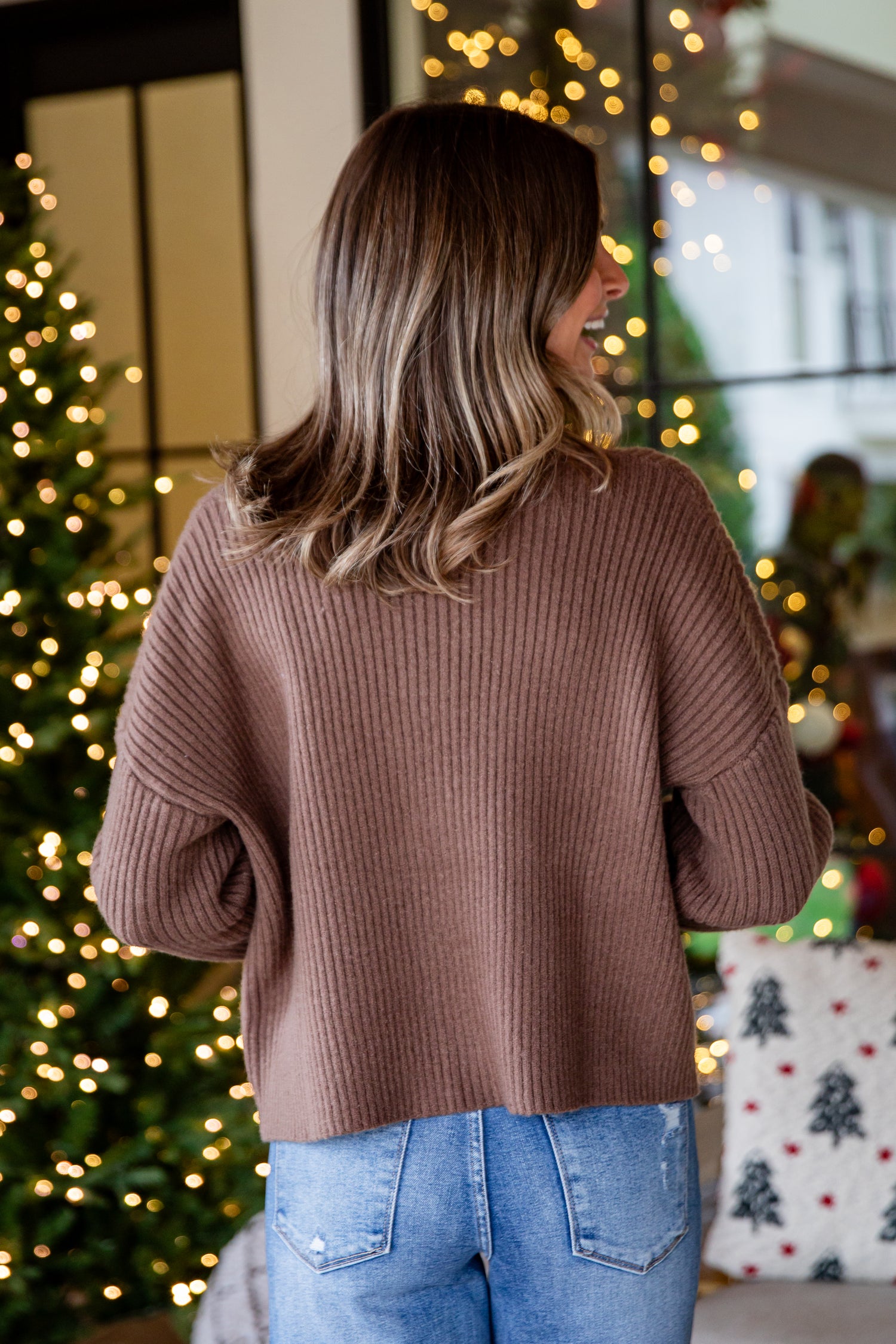 Person wearing a brown sweater and blue jeans standing in front of Christmas decorations.