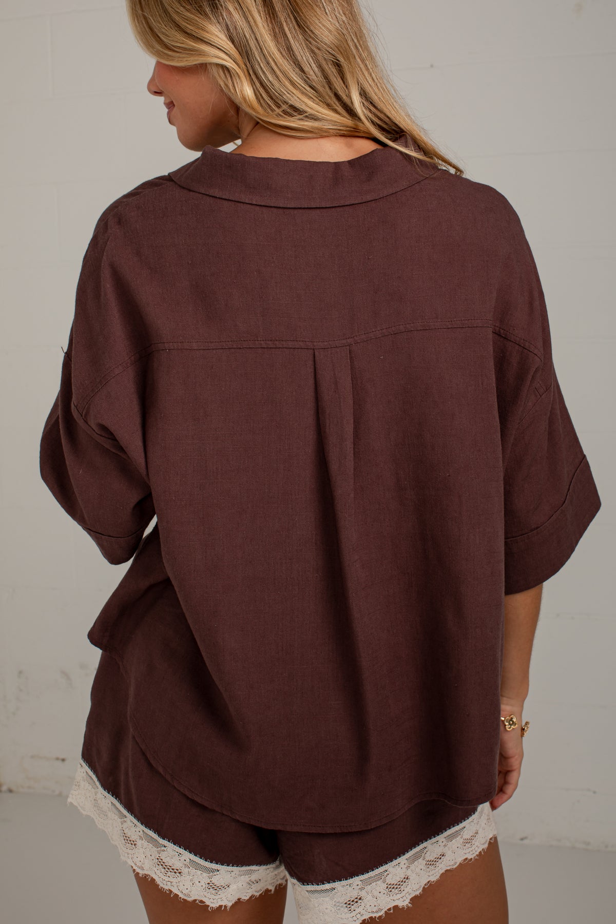 A woman viewed from behind wearing a loose-fitting dark brown short-sleeve top with a pleated back detail and matching shorts featuring white lace trim, set against a plain off-white wall.