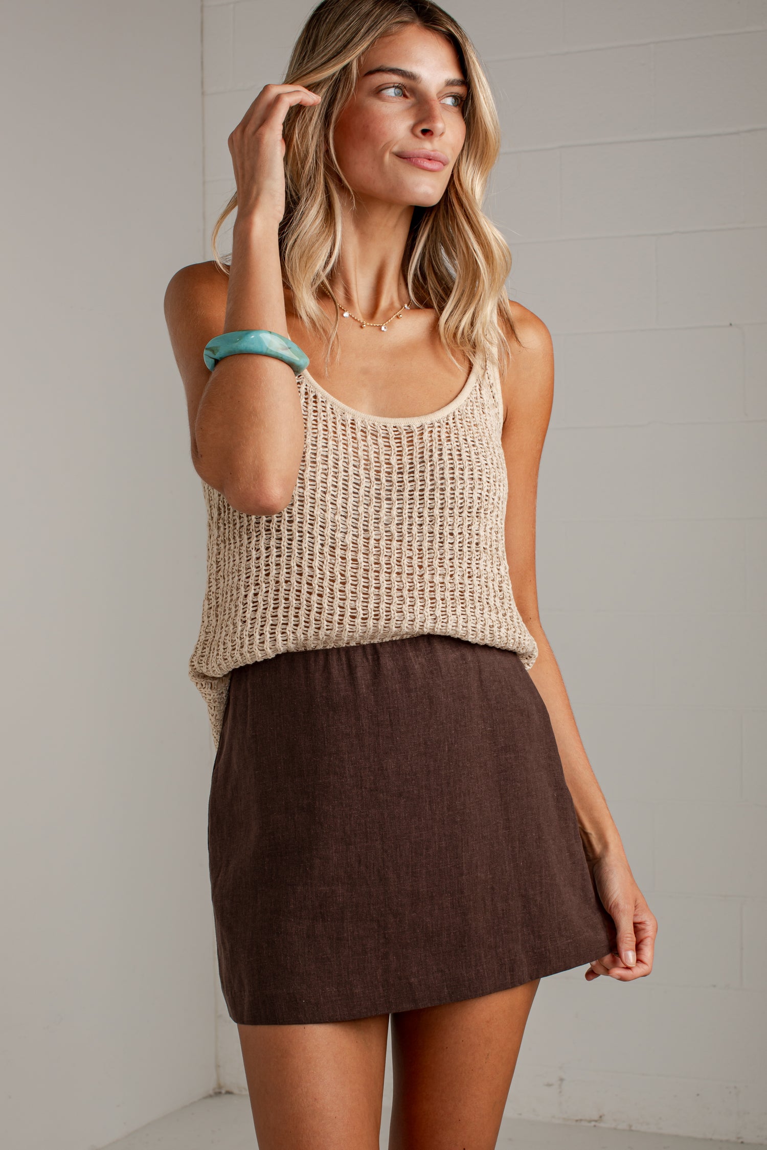 A woman modeling a beige chunky knit tank top and a brown linen mini skirt against a white paneled wall.