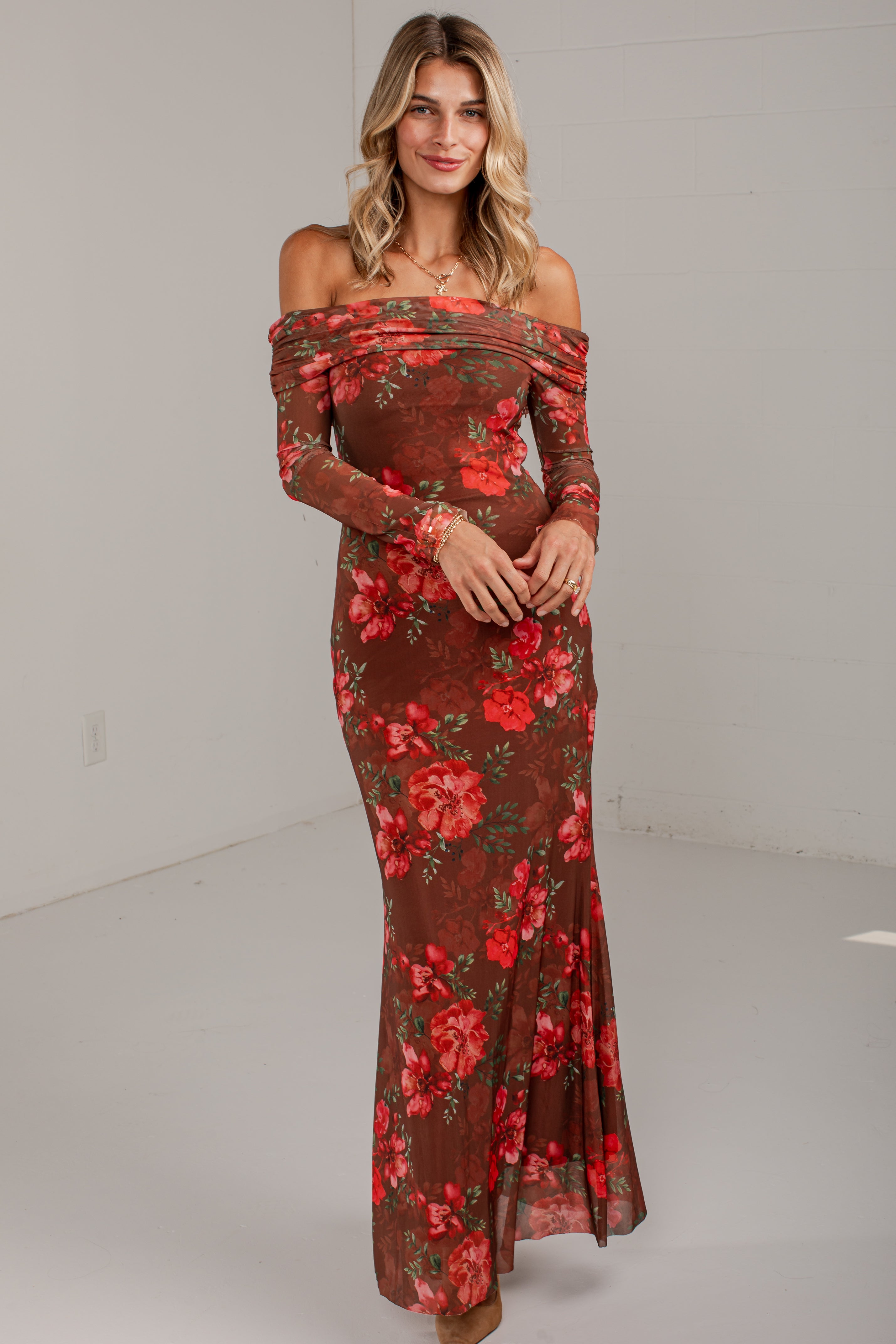 A woman modeling an off-the-shoulder long-sleeve maxi dress with a brown base and a red floral print, standing against a plain light gray studio background.