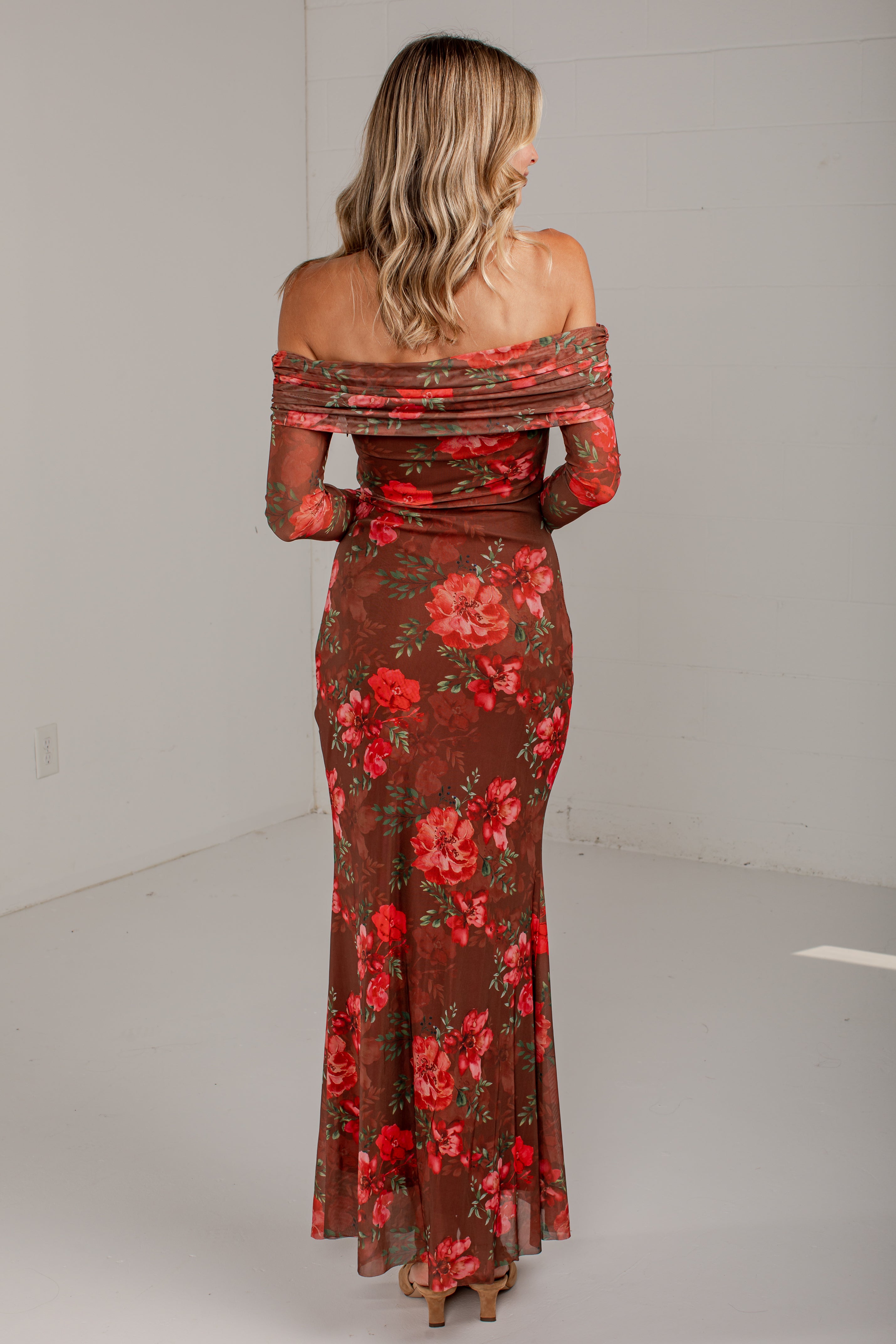 A woman viewed from behind wearing a floor-length off-the-shoulder dress with a sheer brown base and a red floral print, standing in a plain studio setting.