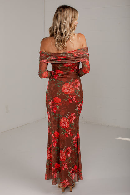 A woman viewed from behind wearing a floor-length off-the-shoulder dress with a sheer brown base and a red floral print, standing in a plain studio setting.