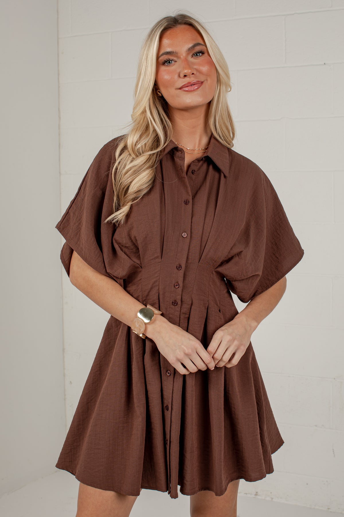 A woman modeling a brown short-sleeve button-front shirt dress against a white brick wall background.