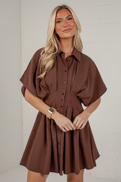 A woman modeling a brown short-sleeve button-front shirt dress against a white brick wall background.