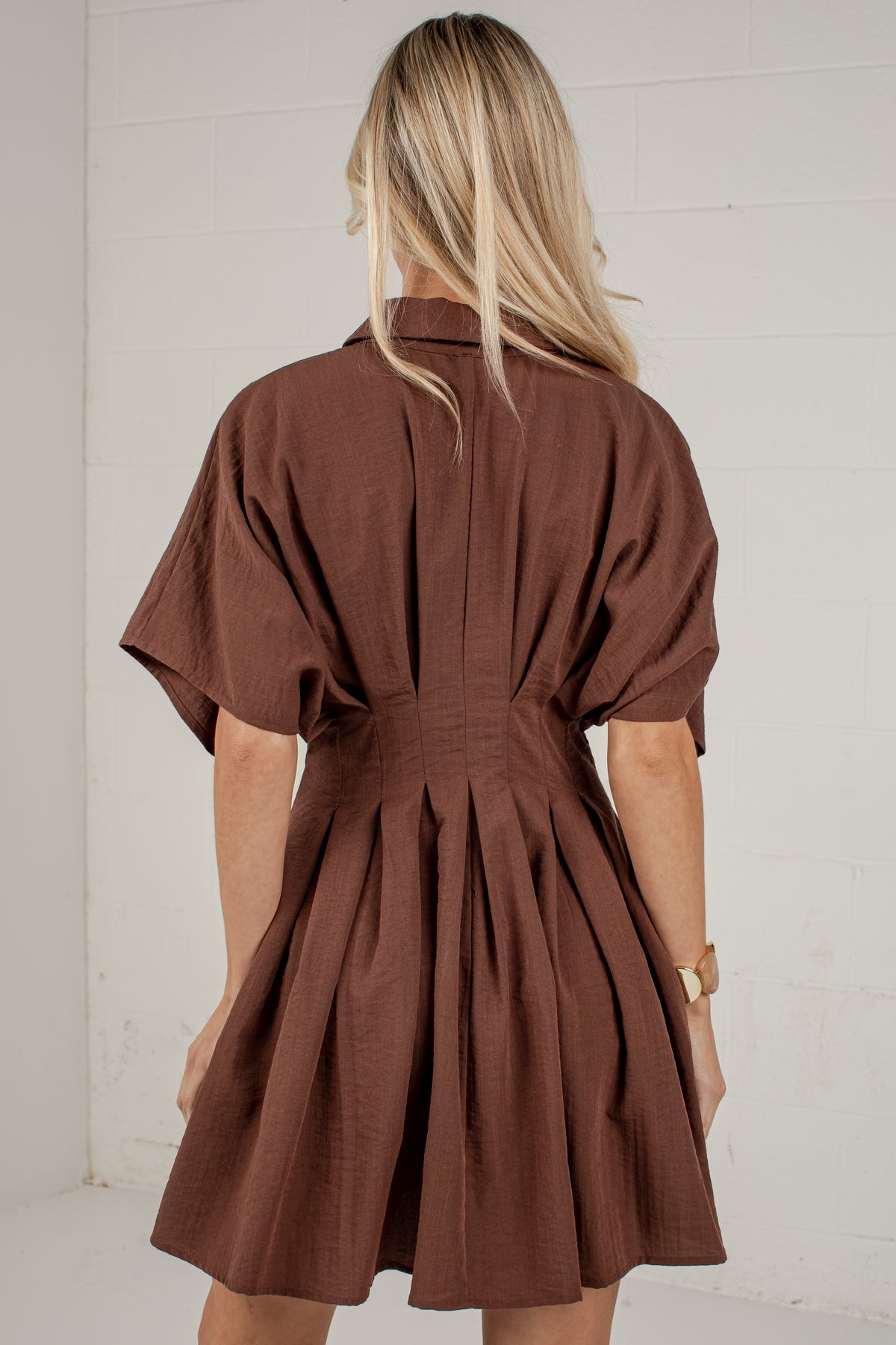 A woman viewed from behind wearing a brown short-sleeve mini dress with a gathered waist and pleated back detail, standing against a white paneled wall.