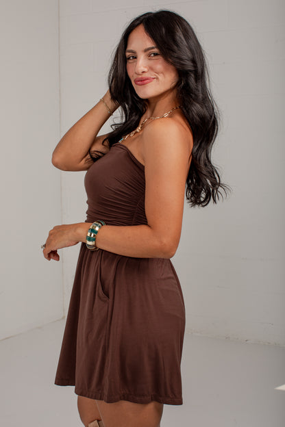 A woman modeling a brown strapless mini dress with a gathered waist and flared skirt, standing against a plain light gray wall.