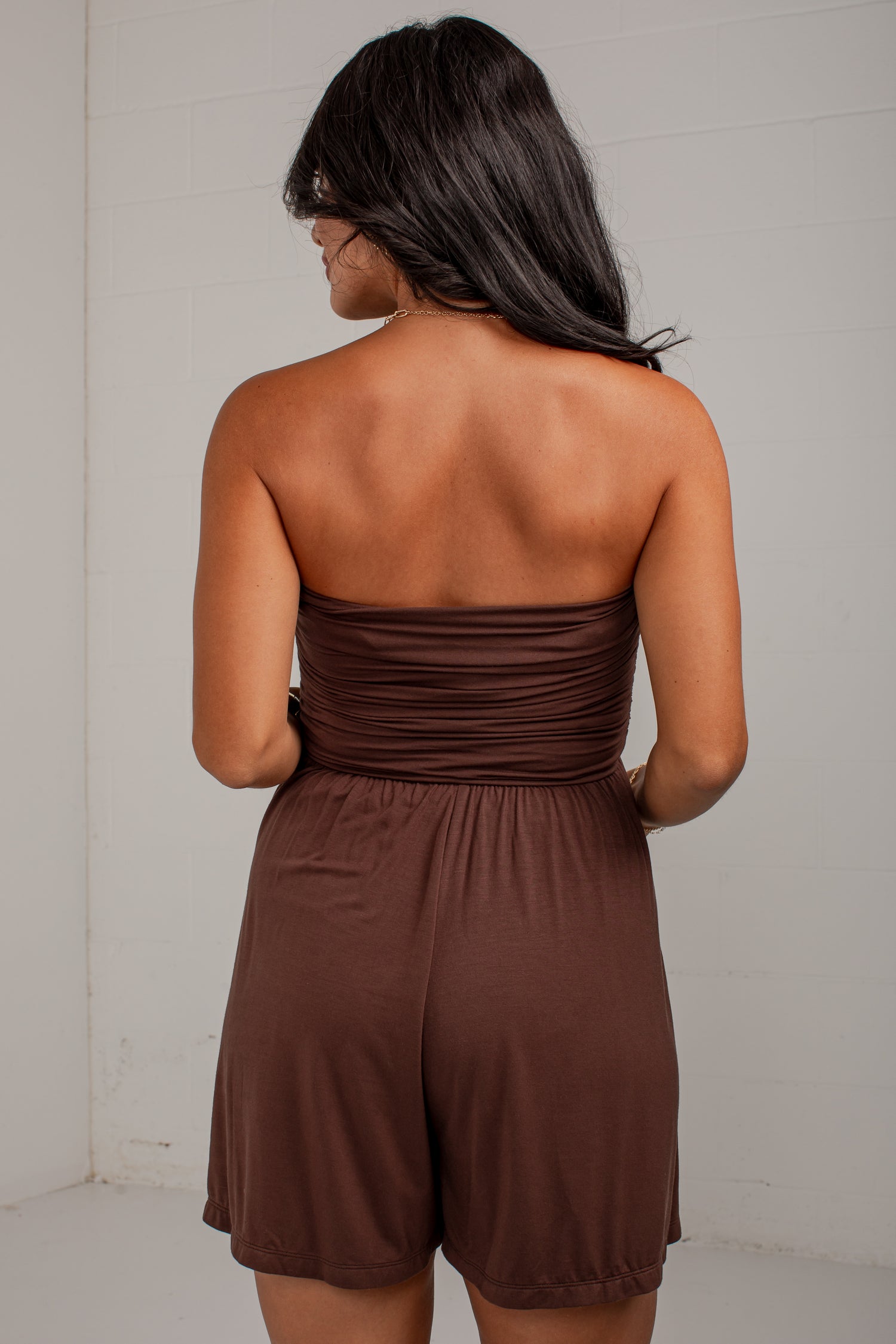 A woman shown from the back wearing a brown strapless romper with a ruched waistband and short legs, standing against a plain light wall.