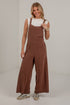 A woman modeling a pair of brown pinstriped wide-leg overall jumpsuits over a white sleeveless top, standing in a white studio setting.