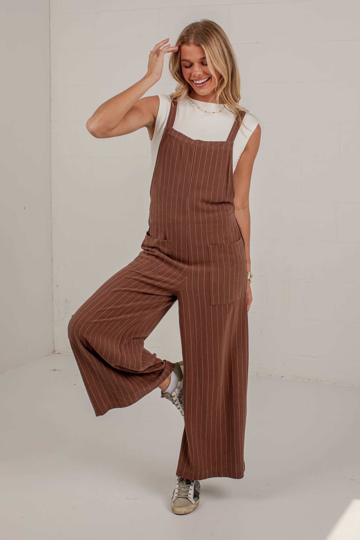 A woman modeling a brown pinstriped wide-leg jumpsuit over a white sleeveless top, standing in a bright studio setting.