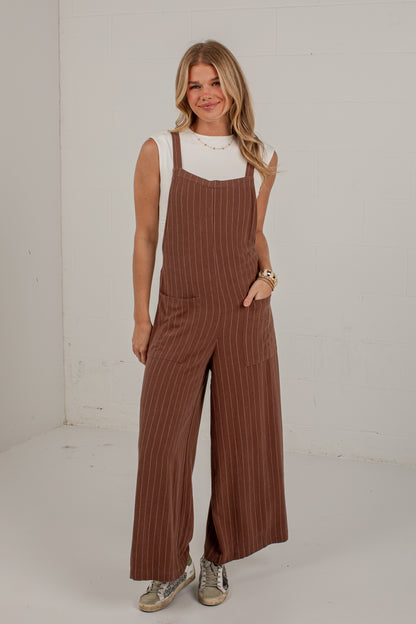 A woman modeling a pair of brown pinstriped wide-leg jumpsuits over a white sleeveless top, standing against a white studio wall.