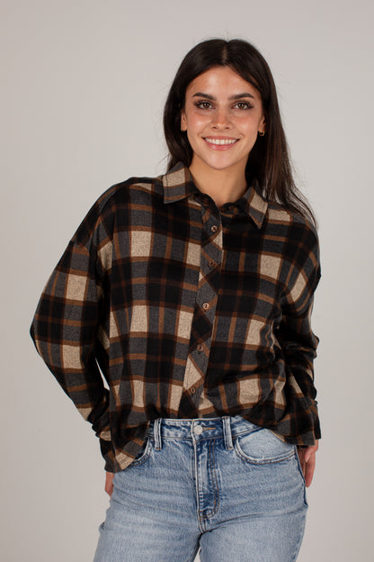 A woman wearing a brown and beige plaid button-down shirt with light blue jeans, standing against a solid light gray background.
