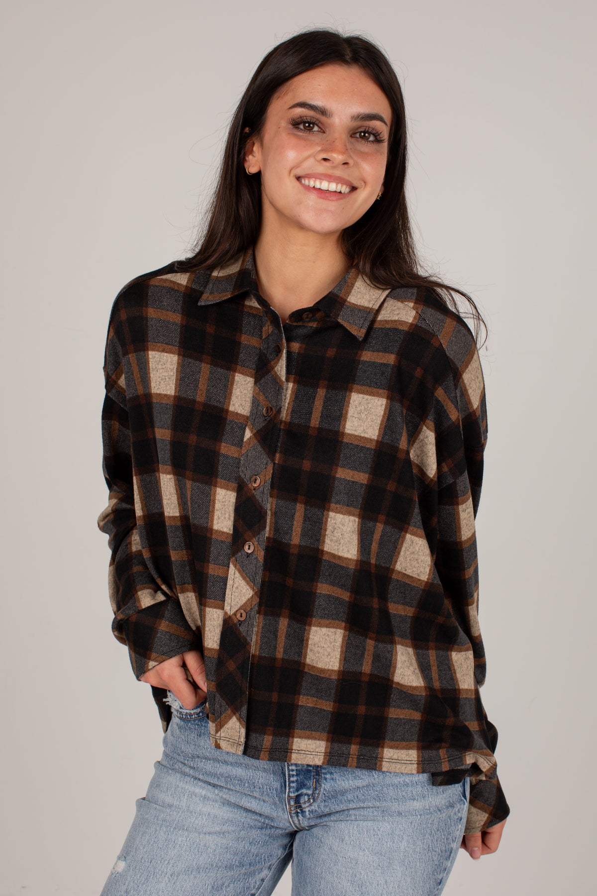 A woman wearing a brown, black, and beige plaid button-up flannel shirt with light blue jeans, standing against a solid light gray background.