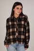 A woman wearing a brown, black, and beige plaid button-down shirt with light blue jeans, photographed against a solid light gray studio background.