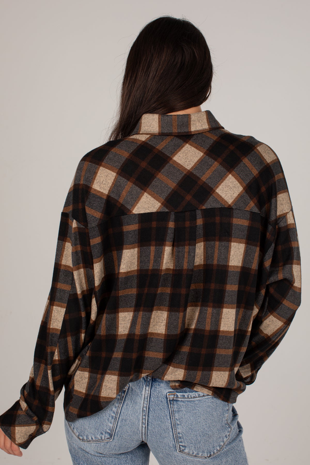 A woman shown from behind wearing a long-sleeve plaid button-up shirt in brown, black, and beige, paired with light blue jeans, against a plain light gray background.