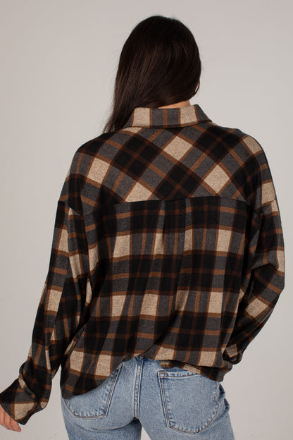 A woman shown from behind wearing a long-sleeve plaid button-up shirt in brown, black, and beige, paired with light blue jeans, against a plain light gray background.
