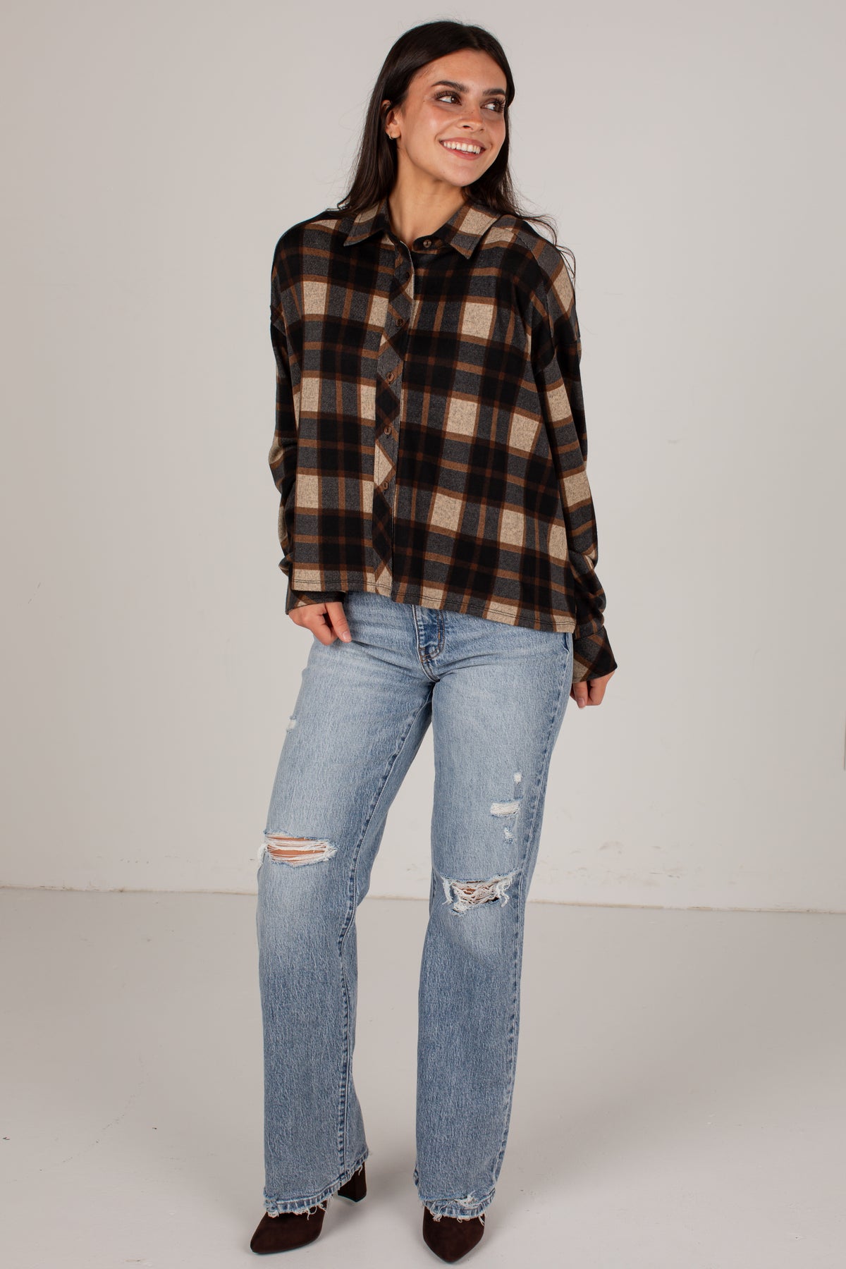 A woman modeling a brown and beige plaid button-down shirt with light-wash distressed jeans, standing against a plain light gray studio background.