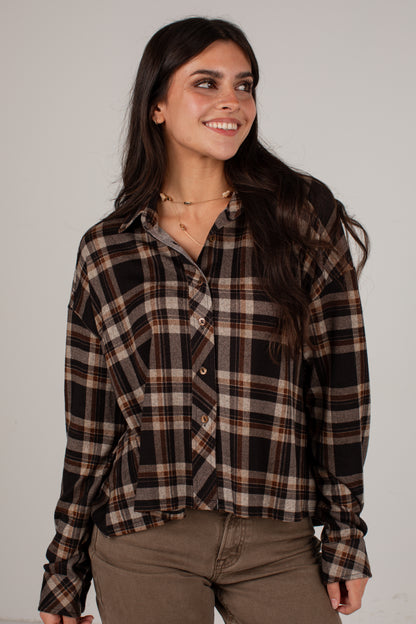 A woman wearing a brown, black, and beige plaid button-up shirt with brown pants, standing against a plain light gray studio background.