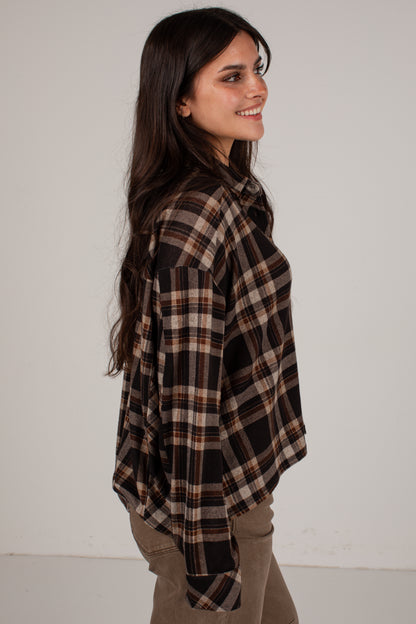 A woman with long dark hair is shown from the side, wearing an oversized brown and black plaid flannel shirt with tan and brown stripes, paired with khaki pants, against a plain off-white background.