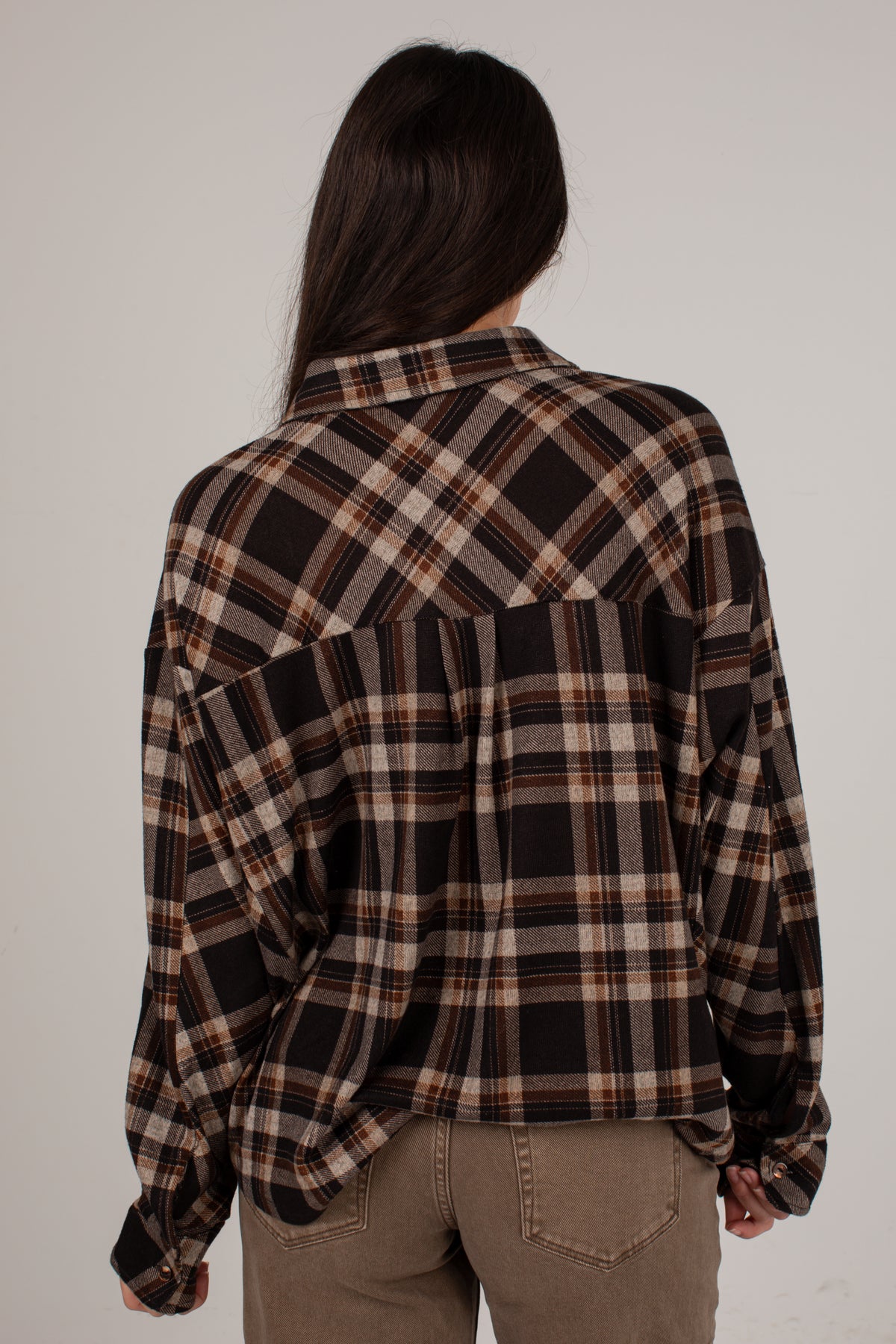 A woman viewed from behind wearing an oversized long-sleeve plaid button-up shirt in brown, black, and beige, paired with khaki pants, against a plain light gray background.