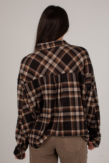 A woman viewed from behind wearing an oversized long-sleeve plaid button-up shirt in brown, black, and beige, paired with khaki pants, against a plain light gray background.
