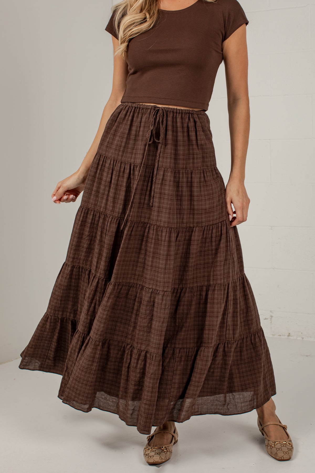 A woman wearing a brown tiered maxi skirt with a subtle plaid pattern, paired with a matching brown short-sleeve crop top and brown flat shoes, standing against a light gray wall.
