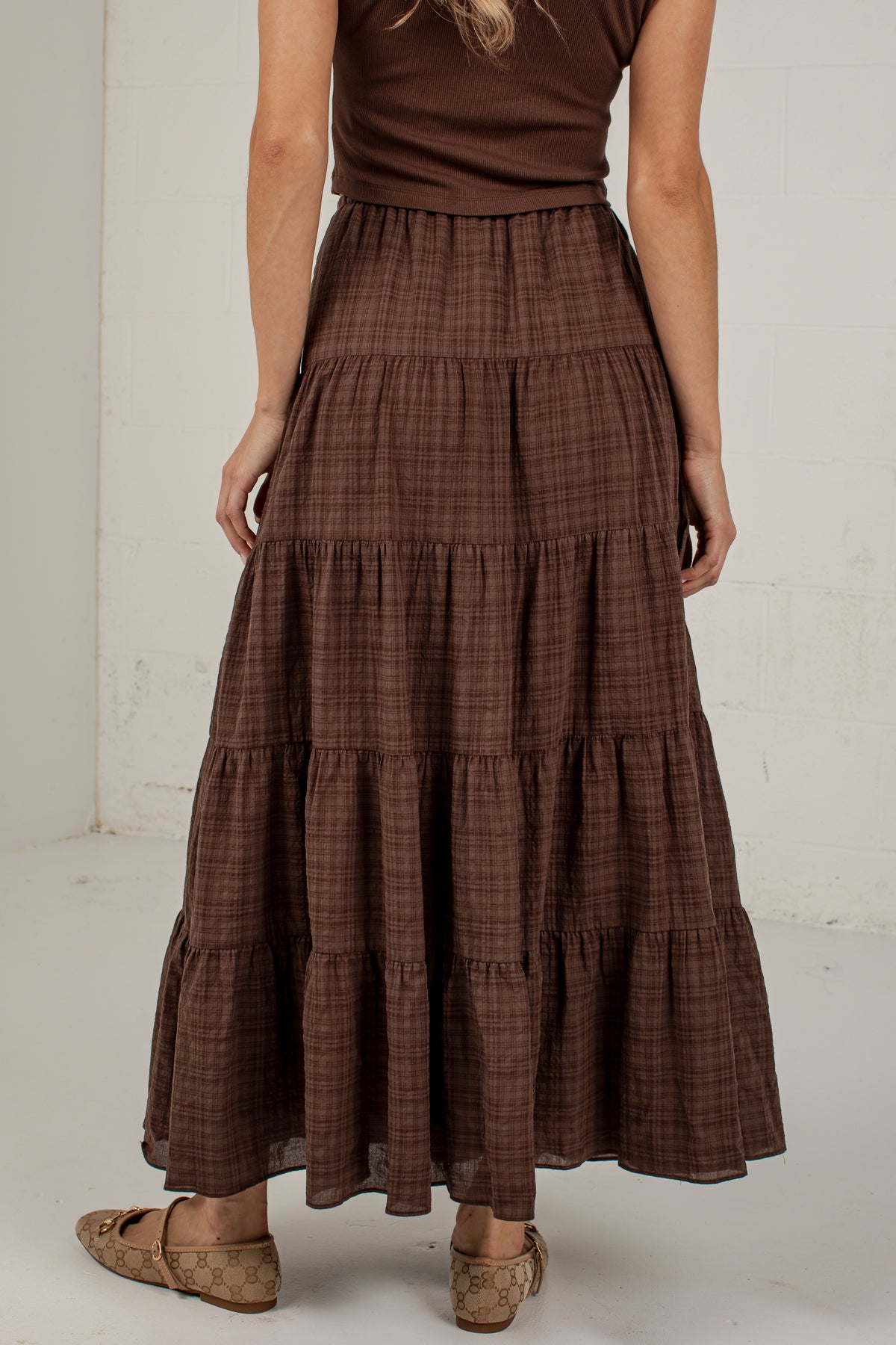 A woman wearing a brown tiered maxi skirt with a subtle plaid pattern, paired with a matching brown sleeveless top and brown flat shoes, standing against a white wall.