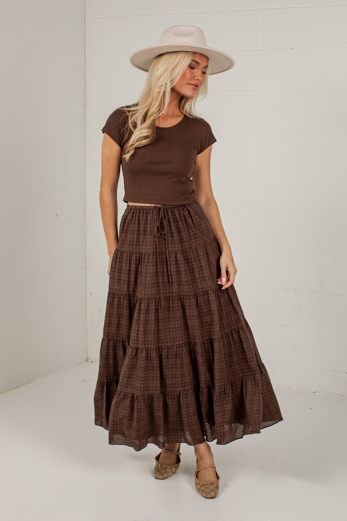 A woman modeling a brown short-sleeve crop top and a matching brown plaid tiered maxi skirt, styled with a beige wide-brim hat and brown flat shoes, set against a white brick wall.