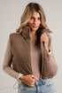 A woman wearing a brown and black houndstooth zip-up vest over a beige long-sleeve top and blue jeans, standing against a white paneled wall.