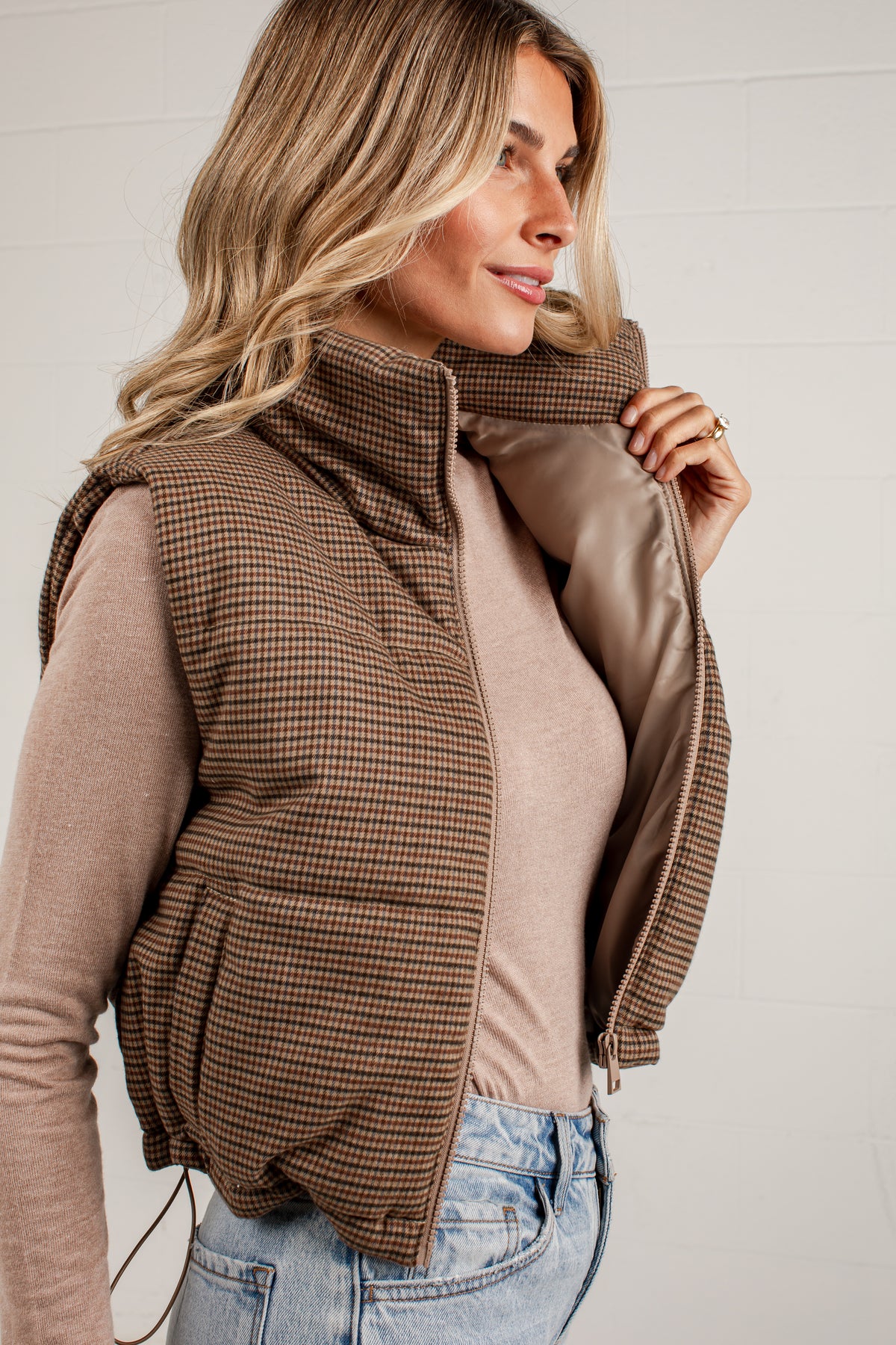 A woman wearing a brown and beige houndstooth puffer vest over a beige long-sleeve top and blue jeans, shown in a close-up profile view against a light gray paneled wall.