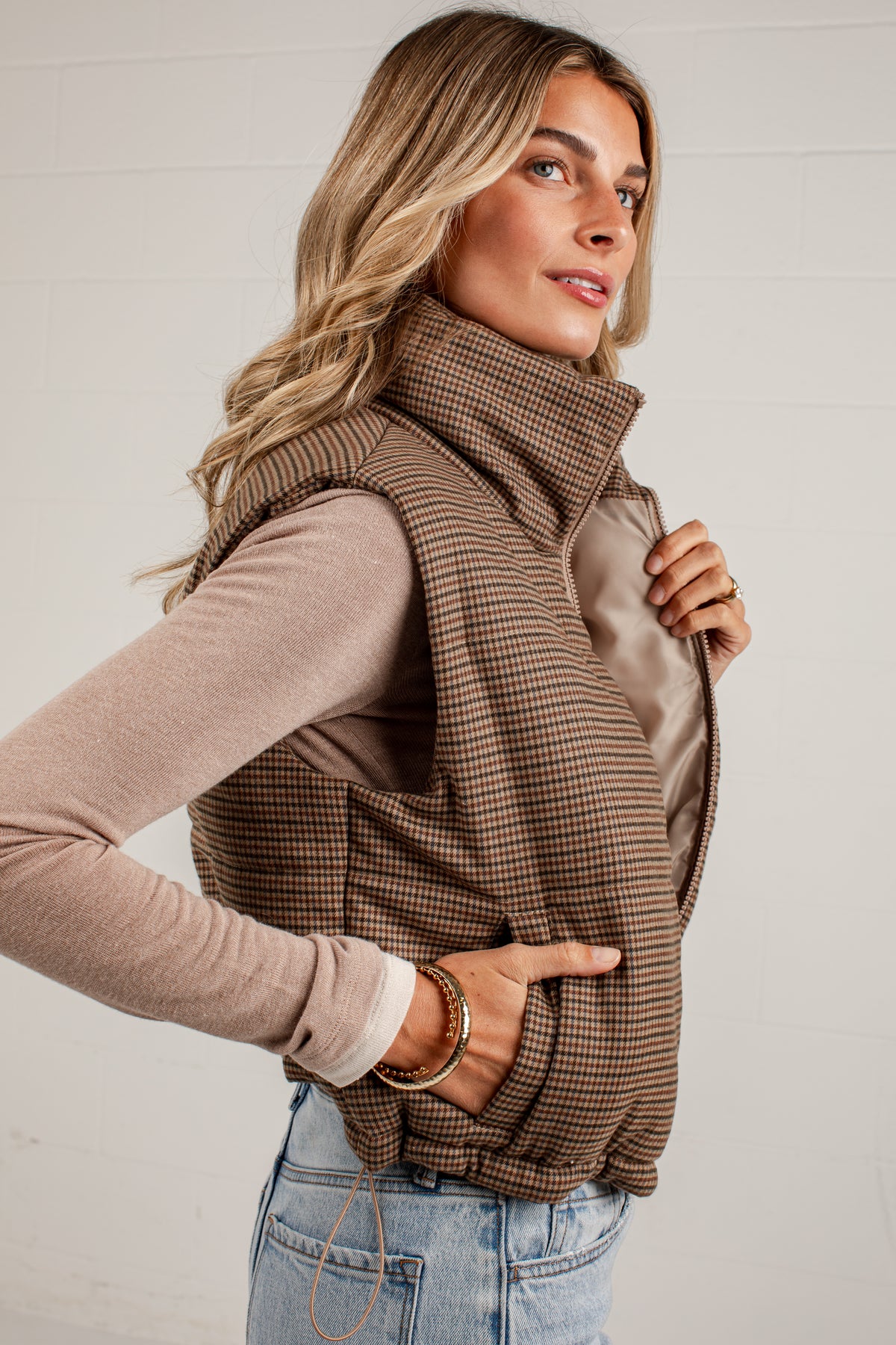 A woman wearing a brown and beige houndstooth zip-up vest over a beige long-sleeve top and light blue jeans, posing against a white paneled wall.