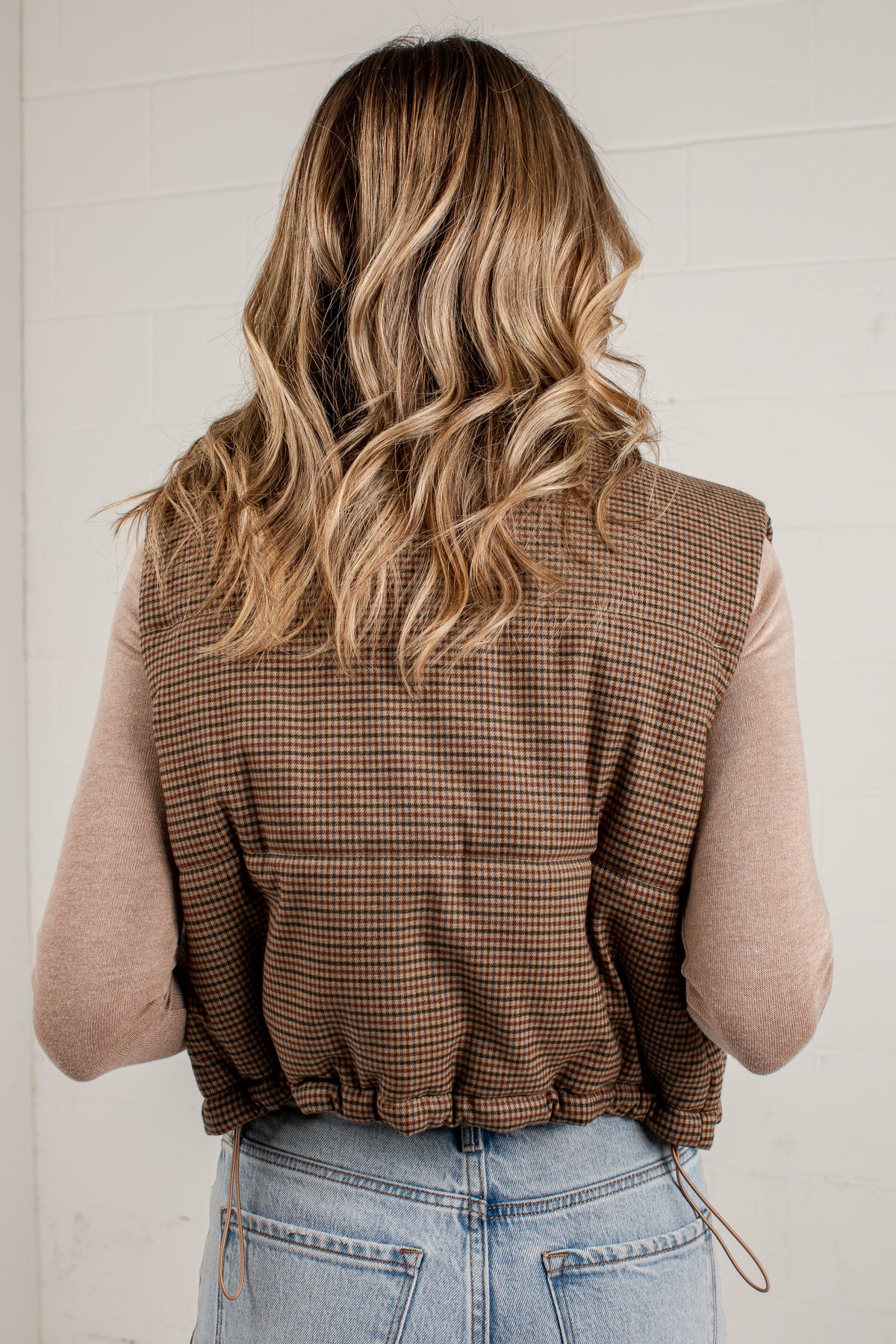 A woman viewed from behind wearing a brown and beige checkered sleeveless vest over a light pink long-sleeve top and light blue jeans, standing against a white brick wall.