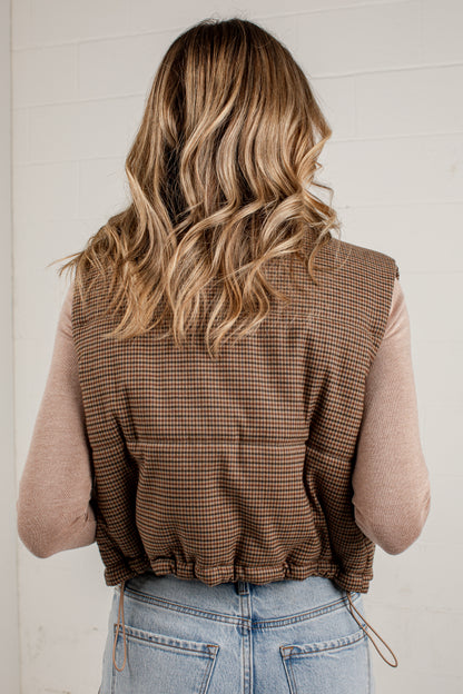A woman viewed from behind wearing a brown and beige checkered sleeveless vest over a light pink long-sleeve top and light blue jeans, standing against a white brick wall.