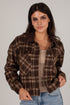A woman wearing a brown and beige plaid button-up flannel shirt jacket over a beige top and blue jeans, photographed against a neutral light gray studio background.