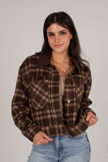 A woman modeling a brown and beige plaid button-up flannel shirt jacket over a beige top and blue jeans, set against a plain light gray studio background.