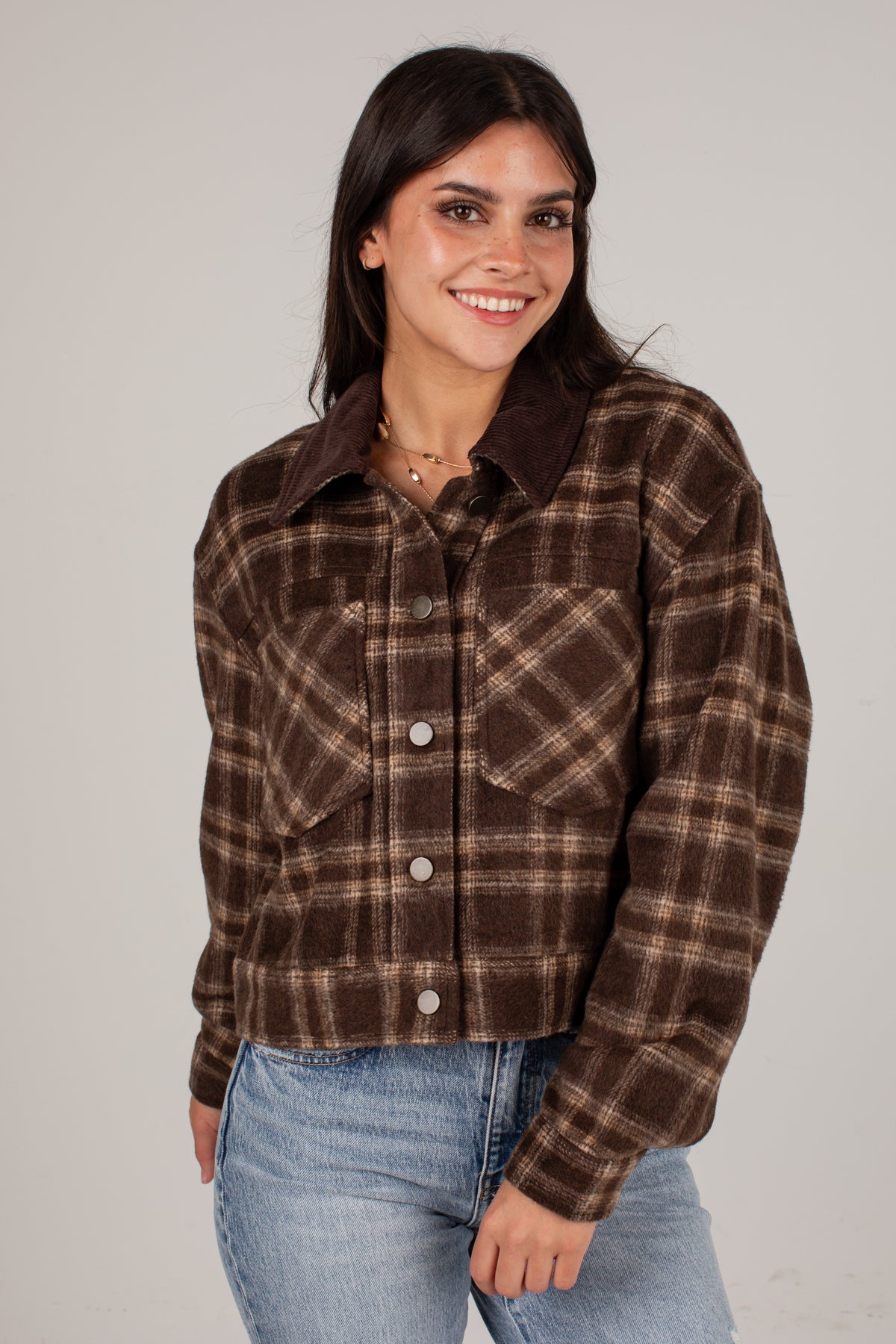 A woman wearing a brown and beige plaid cropped flannel shirt jacket with a solid dark brown collar, paired with light blue jeans, against a plain light gray background.