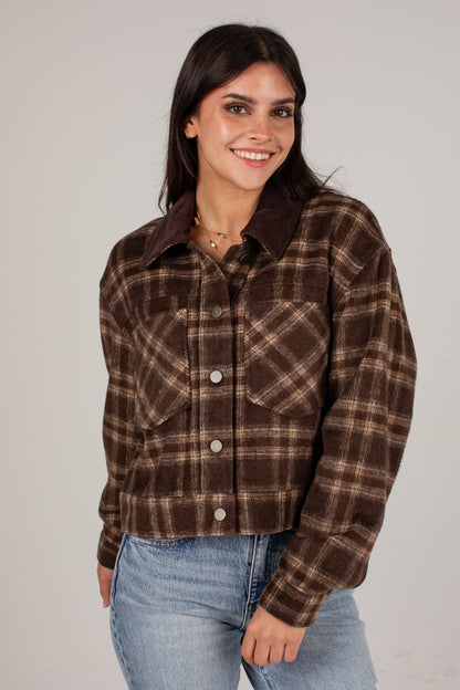 A woman wearing a brown and beige plaid cropped flannel shirt jacket with a solid dark brown collar, paired with light blue jeans, against a plain light gray background.