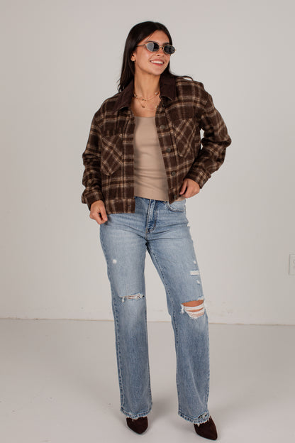 A woman modeling a brown and black plaid flannel shirt jacket over a beige tank top and light-wash distressed jeans, standing against a plain off-white studio background.